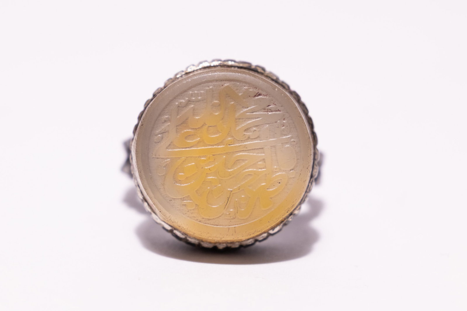 AN ISLAMIC SILVER AND YELLOW AGATE CALLIGRAPHIC INTAGLIO SIGNET RING.: An Islamic signet ring featuring a circular, flat-topped cabochon of translucent pale yellow agate. The stone is intaglio-engraved with Arabic calligraphic script, incorporating the names Allah, Muham