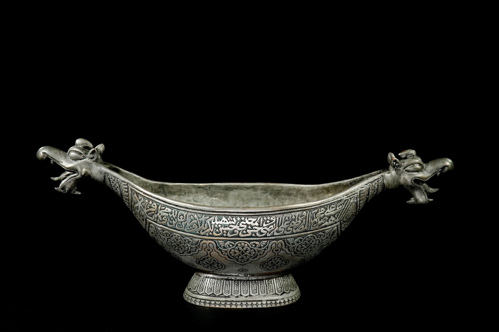 A QAJAR STYLE COPPER KASHKUL WITH DRAGON-HEAD TERMINALS.: A boat-shaped kashkul rising from an oval foot, flanked at each end by sculpted dragon heads with protruding tongues and arched snouts. The exterior is decorated with dense black-infilled motifs, incl