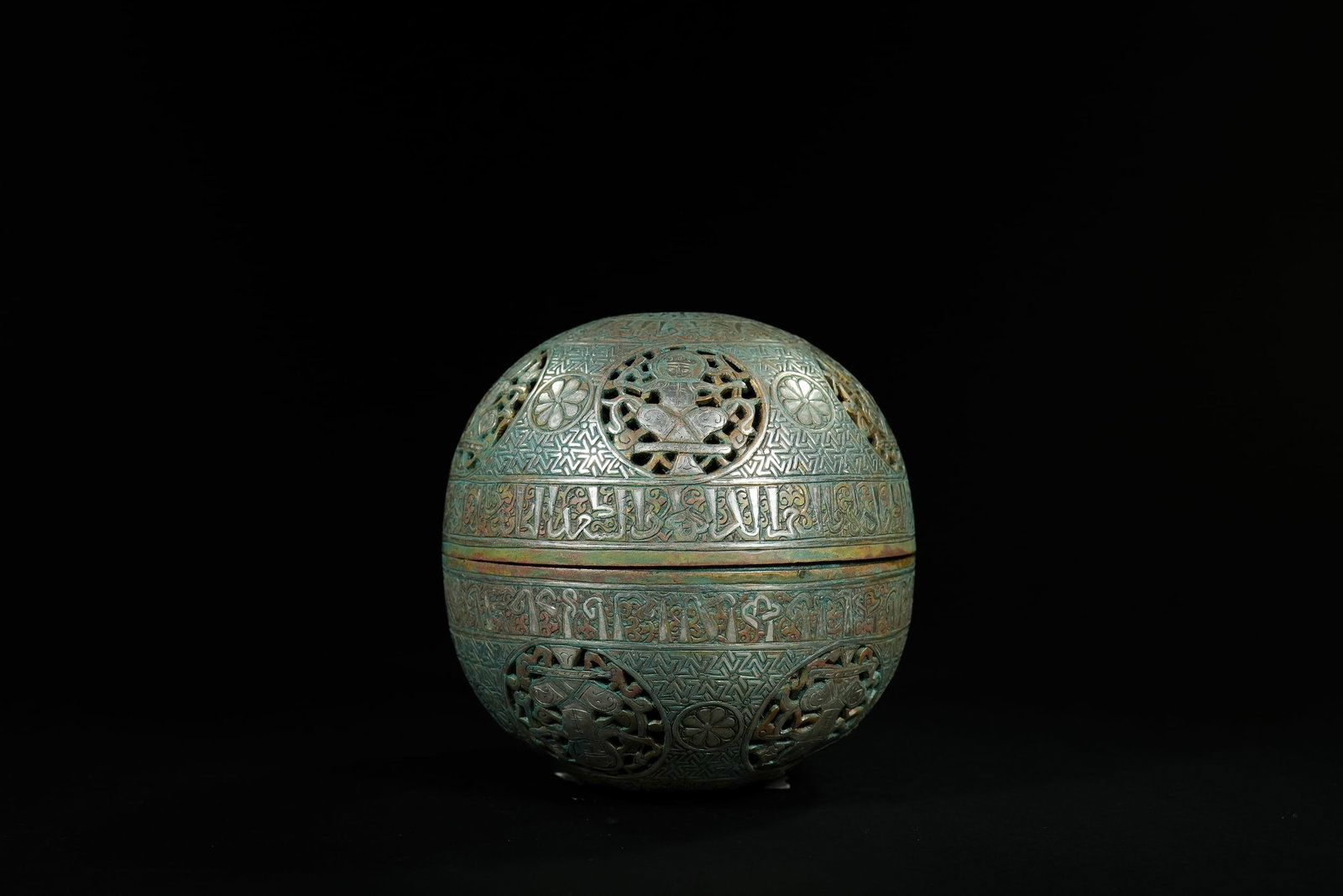 AN ISLAMIC BRASS SILVER-INLAID SPHERICAL LIDDED BOX.: A spherical lidded box composed of brass with silver-inlaid decoration, featuring an all-over design of repeating geometric patterns and circular pierced medallions. The medallions display seated huma