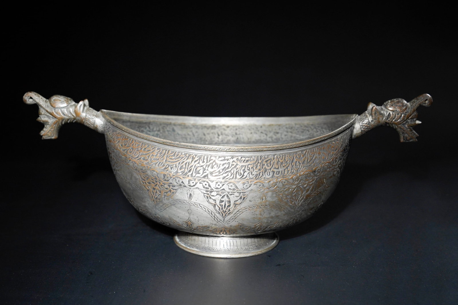 A QAJAR COPPER KASHKUL BEGGAR'S BOWL WITH DRAGON-HEAD TERMINALS, 19TH CENTURY.: An elegant boat-shaped Qajar kashkul finely crafted from copper, standing on a short flared foot. The vessel is richly decorated with bands of intricate thuluth calligraphy, interlaced vegetal scrolls