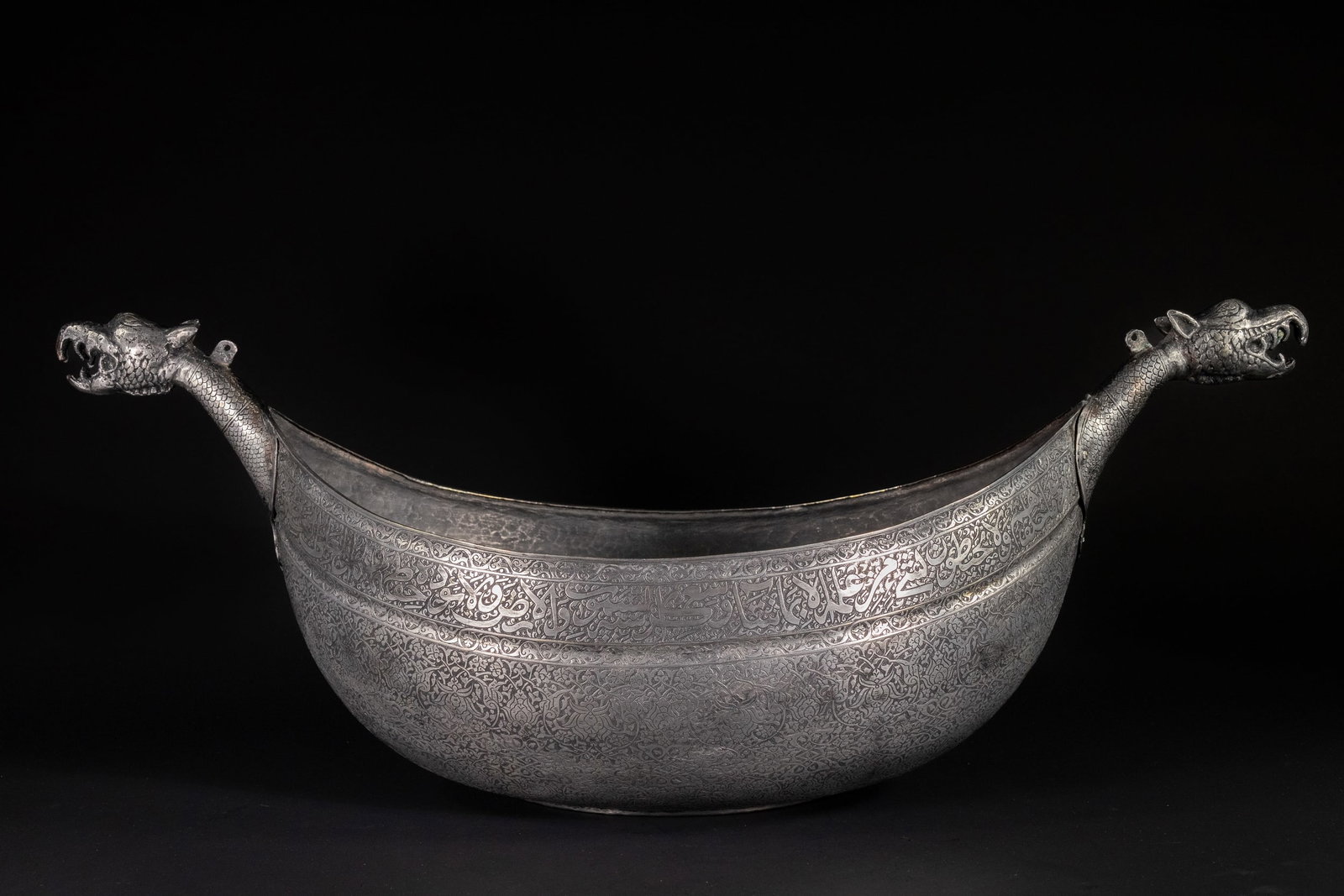 A LARGE TIMURID TINNED COPPER KASHKUL BEGGAR'S BOWL, 15TH CENTURY OR LATER.: A large copper beggarâ€™s bowl kashkul finely engraved with intricate vegetal scrollwork across its body and neck. The upper band features an extensive Arabic inscription in thuluth script, li