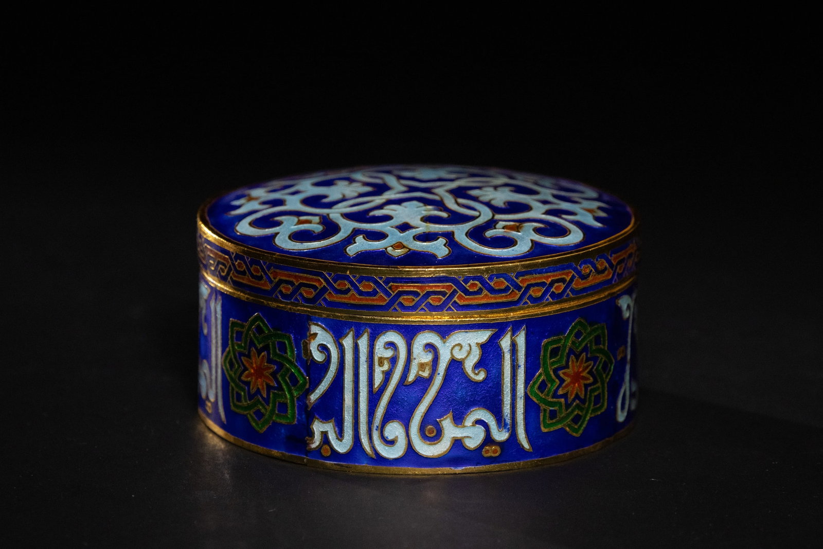 A MAMLUK STYLE ENAMELED CIRCULAR BOX WITH ARABIC CALLIGRAPHY.: A vibrant circular lidded box featuring deep cobalt-blue enamel ground with striking turquoise and white scrollwork on the lid. The body is encircled by alternating eight-pointed star medallions and s