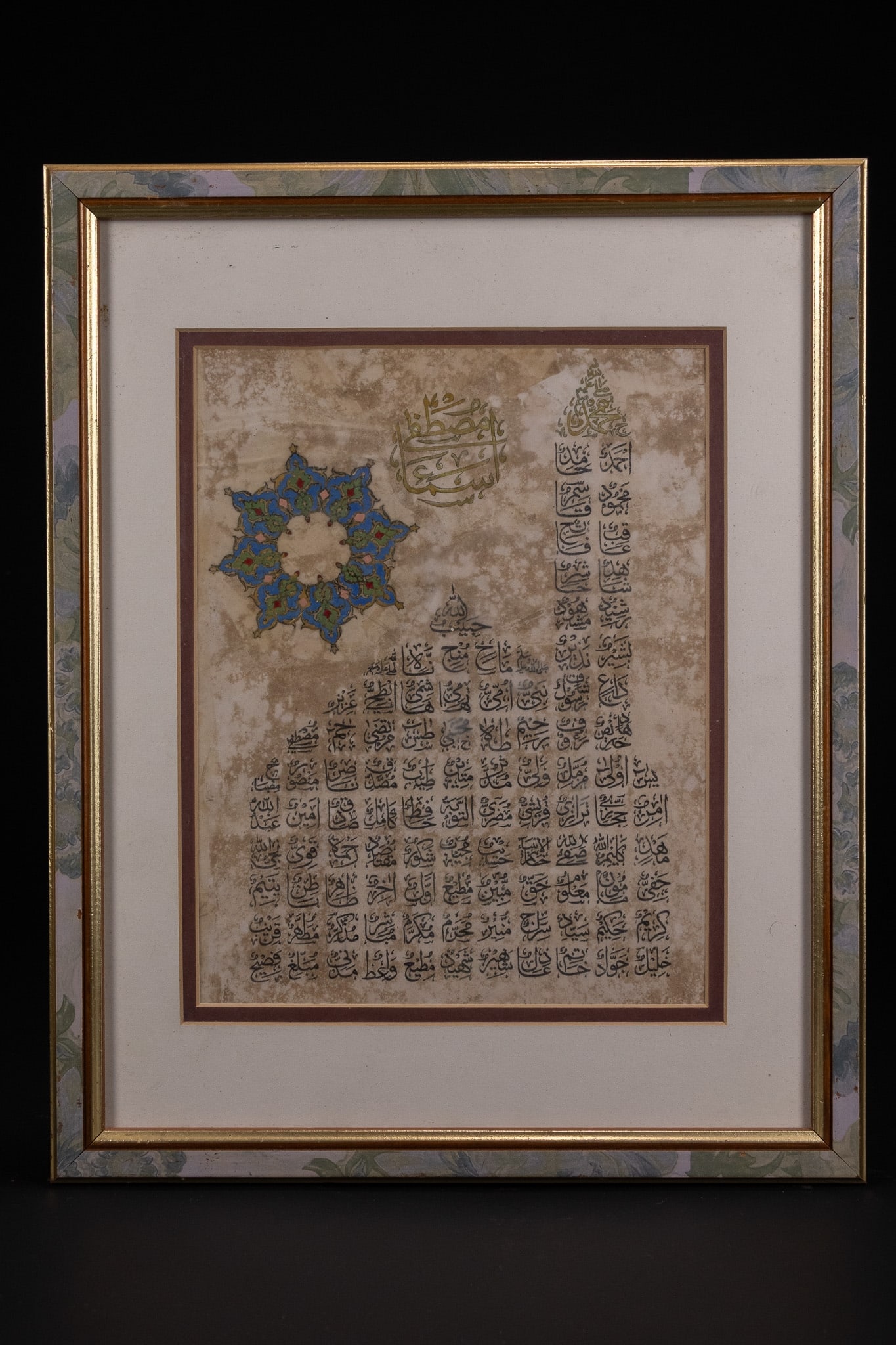 AN ISLAMIC CALLIGRAPHIC PANEL WITH ILLUMINATED ROSETTE, INK AND POLYCHROME ON PAPER.: A refined calligraphic composition featuring densely arranged Arabic script in structured blocks, executed in black ink across a softly mottled ground. The upper section is adorned with a prominent go