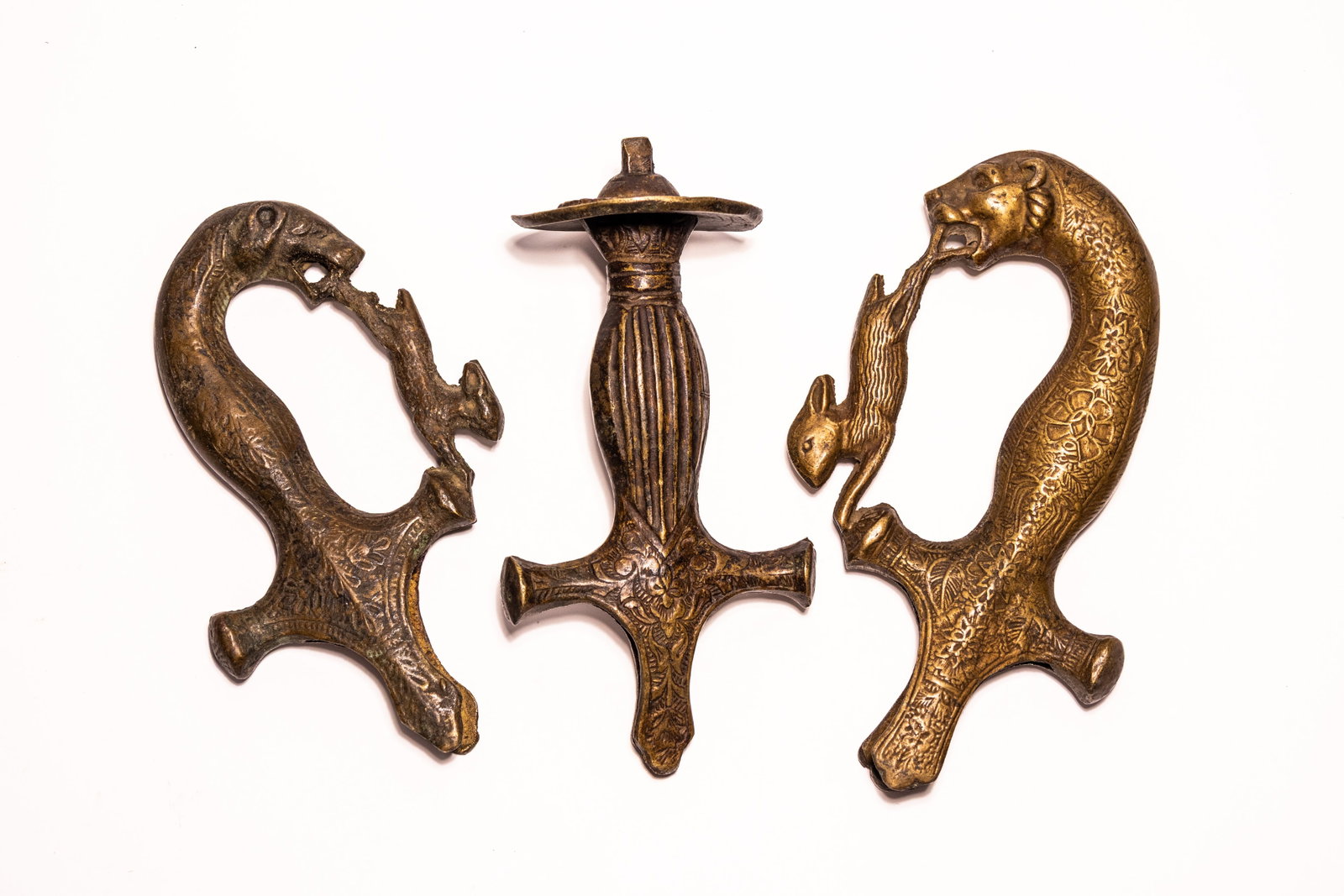 A TRIO OF INDIAN CAST BRASS SWORD HILTS, 19TH CENTURY.: This lot comprises three Indian cast copper alloy sword hilts, featuring one traditional disc-pommel tulwar hilt and two decorative zoomorphic hilts. The central tulwar hilt is designed with a large, 