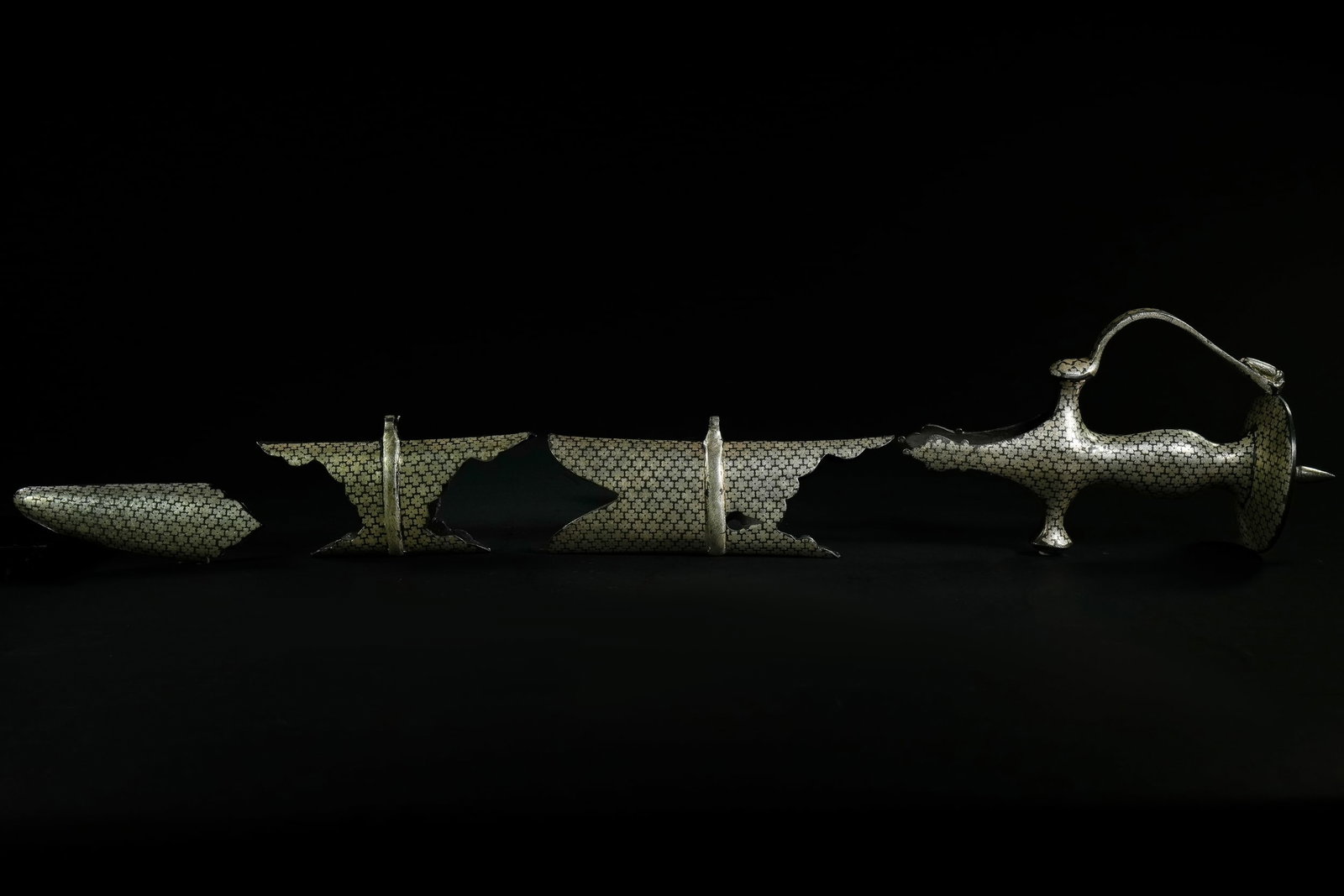 AN INDIAN STEEL TALWAR WITH SILVER-INLAID FLORAL MOTIFS, 20TH CENTURY.: A fragmented Indian steel talwar featuring an elegant surface fully decorated with repeating silver-inlaid floral motifs across the hilt, guard, and remaining blade sections. The hilt displays a bulbo