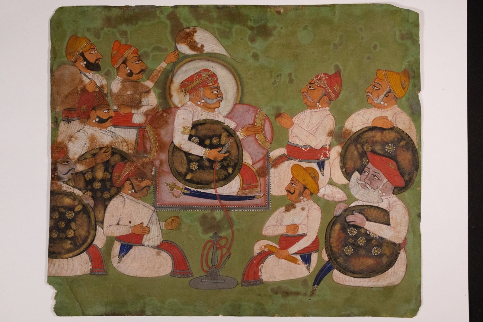 AN INDIAN MINIATURE DEPICTING A MUGHAL NOBLE IN COURTLY ASSEMBLY WITH ATTENDANTS, LATE 18TH CENTURY.: A detailed Mughal miniature painting depicting a nobleman seated on a pink bolster throne, flanked by attendants and courtiers in a ceremonial durbar setting. The central figure, adorned in white robe