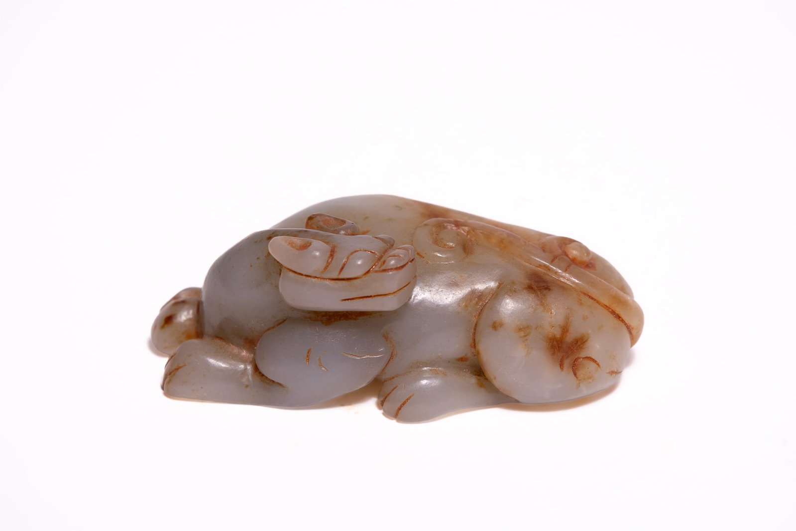 A CHINESE PALE GREEN AND RUSSET JADE CARVING OF A MYTHICAL BEAST, POSSIBLY MING OR QING DYNASTY.: A Chinese carved nephrite jade figure of a mythical beast, depicted in a recumbent position with its head turned back over its shoulder. The stone is of a pale celadon and greyish-white colour, incorp