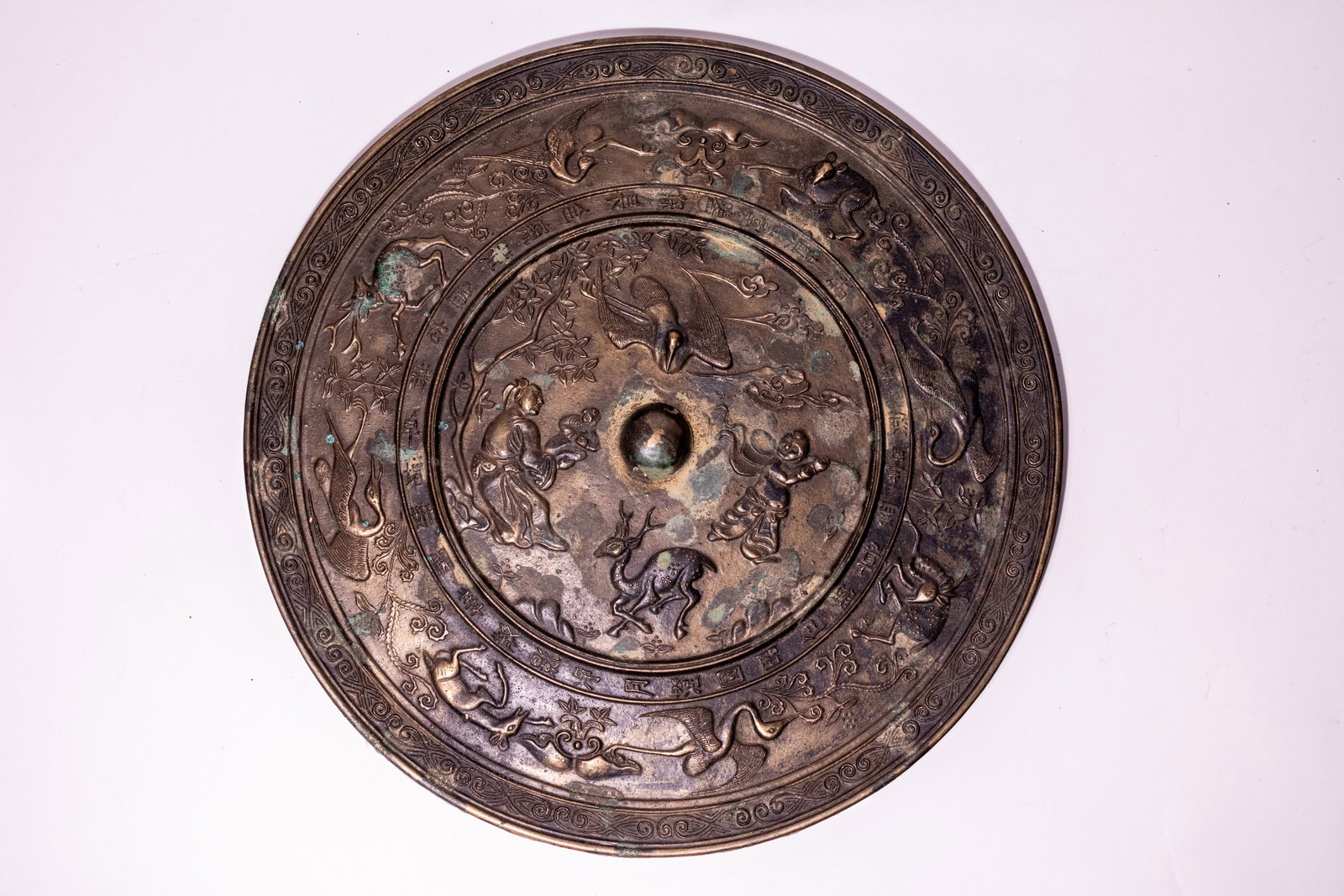 A LARGE CHINESE TANG DYNASTY STYLE BRONZE MIRROR WITH FIGURES, DEER, AND CRANES.: This Chinese cast bronze mirror of circular form features high relief decoration on the reverse side. The central field is arranged around a pierced domed knop, depicting a seated sage or immortal fig
