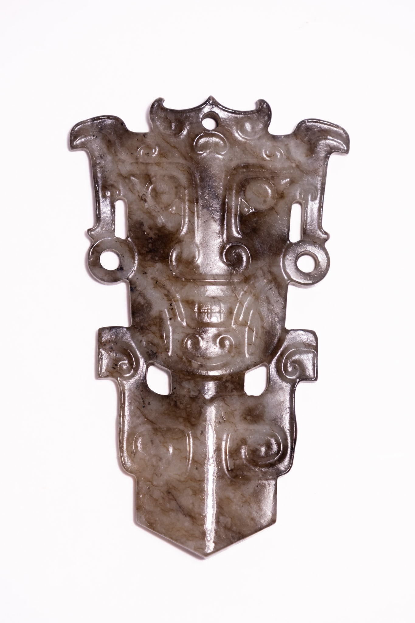 A CHINESE CARVED ARCHAIC STYLE MOTTLED JADE TAOTIE MASK PLAQUE, 18TH-19TH CENTURY.: A symmetrical carved plaque produced in the archaic style from mottled celadon and black jade. The piece features a central taotie monster mask motif with prominent swirling eyes, a flattened nose, an