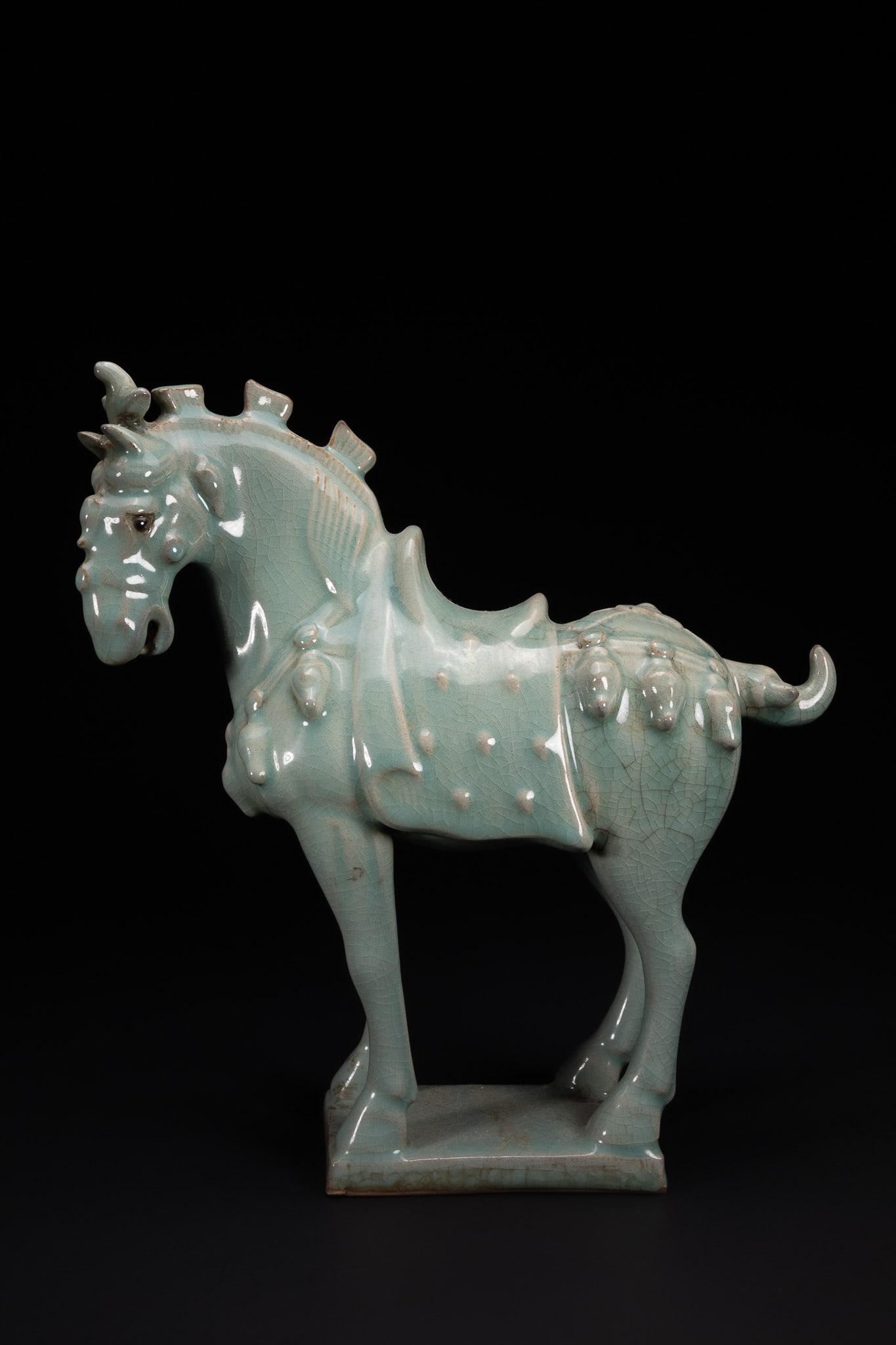 A CHINESE CELADON-GLAZED MODEL OF A HORSE.: A Chinese ceramic model of a horse, modelled standing on a rectangular base with slender legs and an elongated body. The horse features a stylised head with pronounced eyes, a slightly open mouth, and
