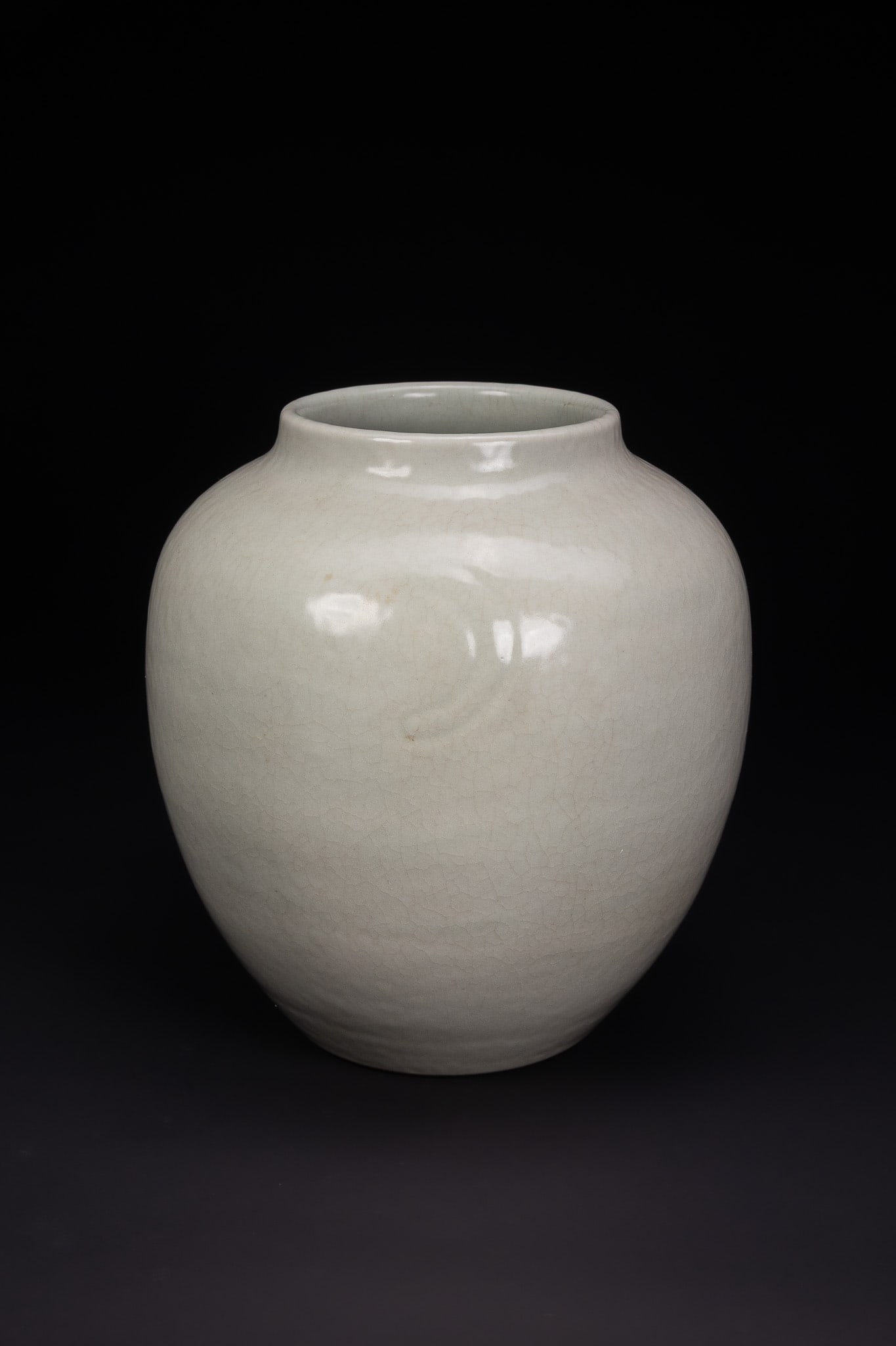 A CHINESE CELADON-GLAZED CRACKLED JAR, QING DYNASTY.: This large porcelain jar is of globular form, featuring a well-rounded body that tapers slightly towards a short, wide neck and a rounded rim. The vessel is enveloped in a thick, pale celadon glaze wi