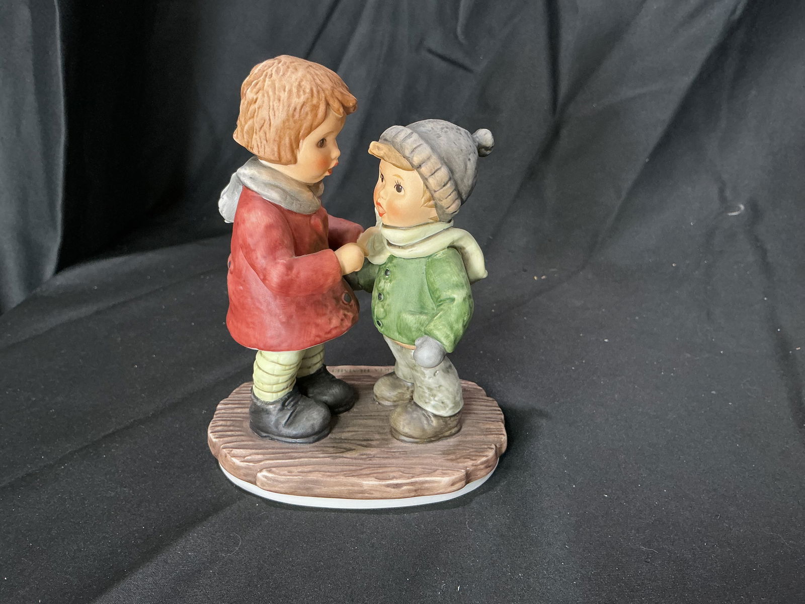 Berta Hummel "Snug and Warm" Figurine by Goebel: Berta Hummel figurine titled "Snug and Warm," depicting two children standing close together in winter clothing, sharing a tender moment. One child wears a red coat and scarf, while the other is dress