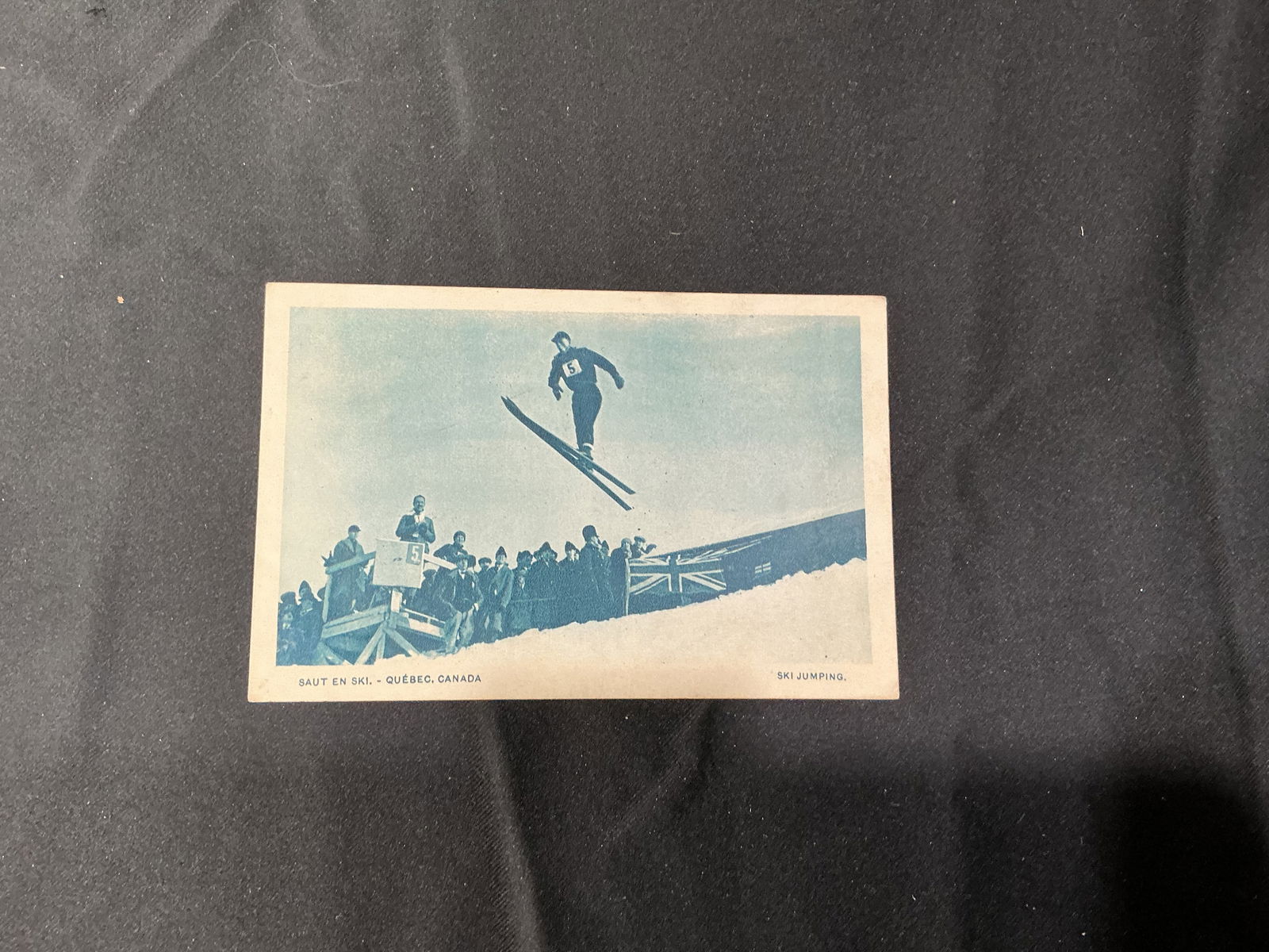 Vintage 'Saut en Ski’ Quabec, Canada' Ski Jumping Postcard: Original vintage postcard titled 'Saut en Ski’ Quabec, Canada' (Ski Jumping) depicting a ski jumper midair above a cheering crowd and flags at a winter sporting event. The image captures the early 2