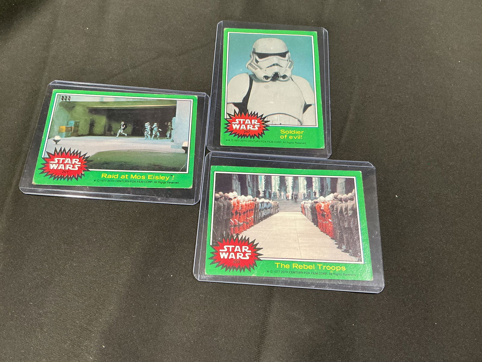 Three vintage 1977 Star Wars trading cards (green border): Group of three vintage Star Wars trading cards with classic green border designs, including: 'Raid at Mos Eisley!', 'Soldier of evil!' (Stormtrooper), and 'The Rebel Troops'. Cards are housed in