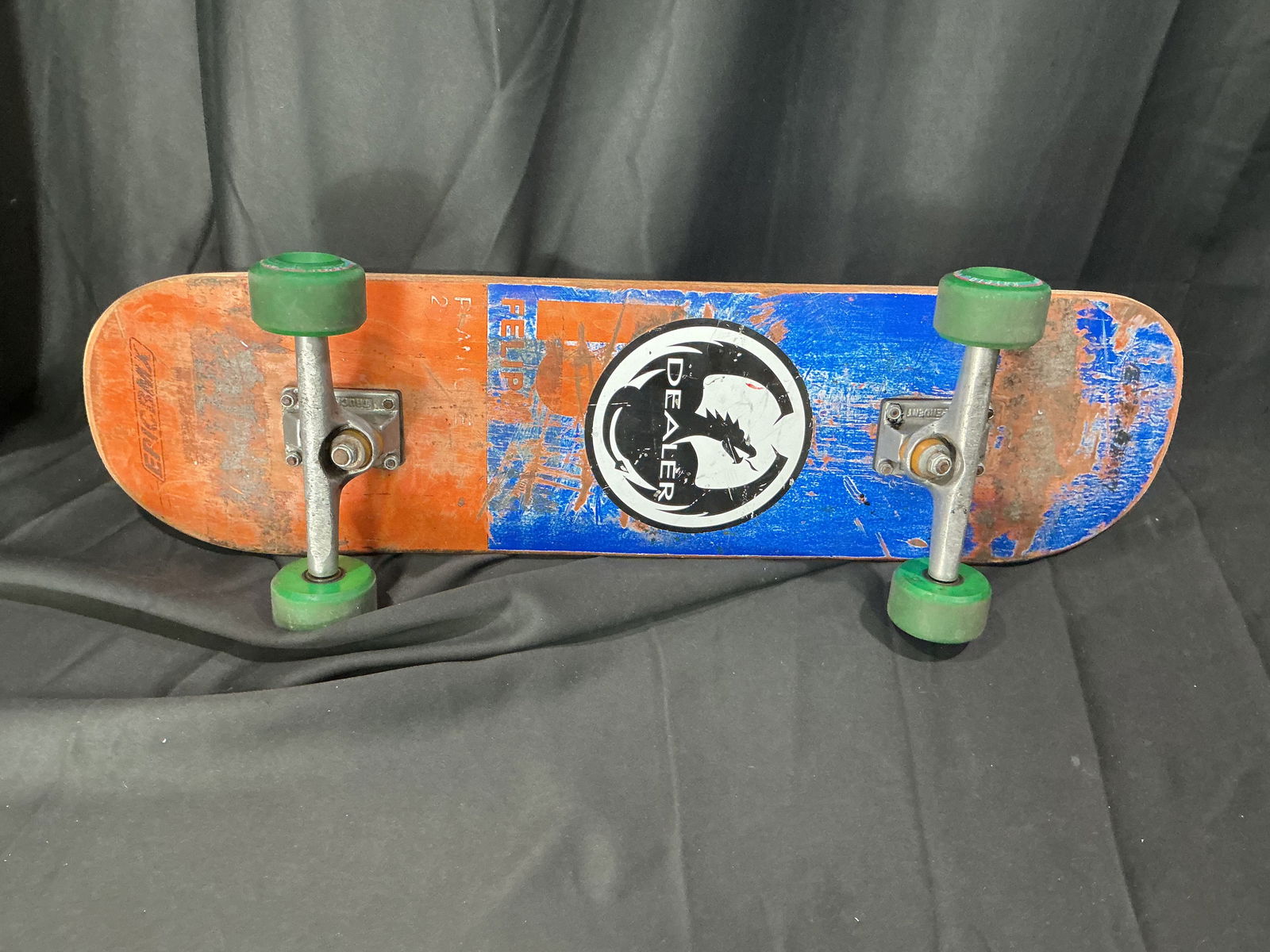 Vintage Dealer-branded complete skateboard with green wheels: Complete skateboard featuring a Dealer-branded deck with orange and blue painted graphic underside. Fitted with metal trucks and green wheels. The top retains applied grip tape. Deck branding and hard