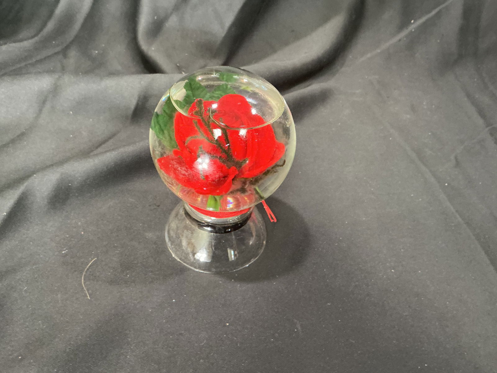Glass dome paperweight with artificial rose: Decorative clear glass dome paperweight containing a red artificial rose with green leaves. Mounted on a matching clear glass pedestal base. Intended as a decorative accent or keepsake display.
