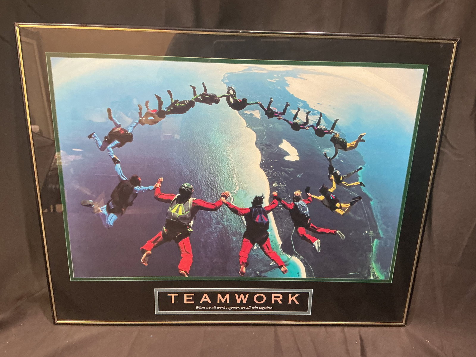 Teamwork Motivational Poster Skydivers Forming Circle (Successories-style): Iconic aerial photograph of a large group of skydivers holding hands in perfect circular formation high above a coastal landscape. Bold lettering reads TEAMWORK with the quote When we all work