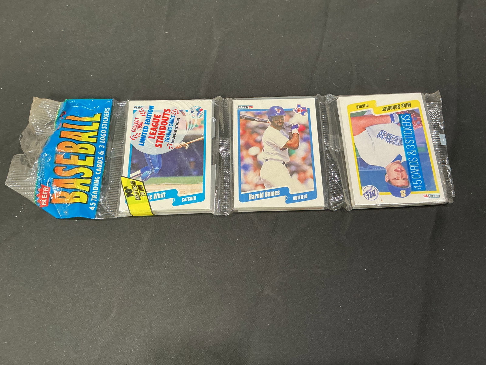 1990 Fleer Baseball Wax Pack Featuring Harold Baines & Mike Schooler: Sealed 1990 Fleer baseball wax pack featuring visible Harold Baines Texas Rangers card and Mike Schooler Seattle Mariners card on outer stack. Includes logo stickers and league standouts insert