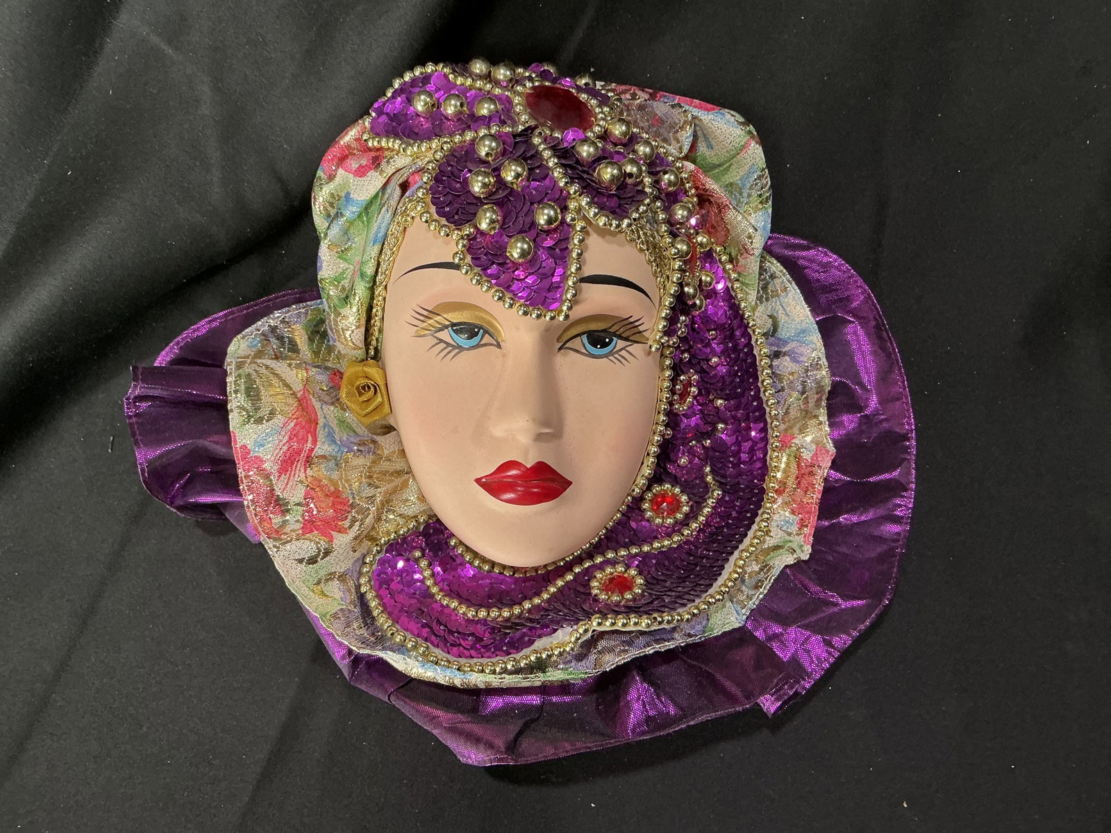 Decorative Venetian-Style Masquerade Mask with Purple Sequin Headdress: Making another appearance, this decorative Venetian-style masquerade mask features a serene female face framed by an ornate purple sequin and gold bead headdress. Layered floral-patterned fabric and