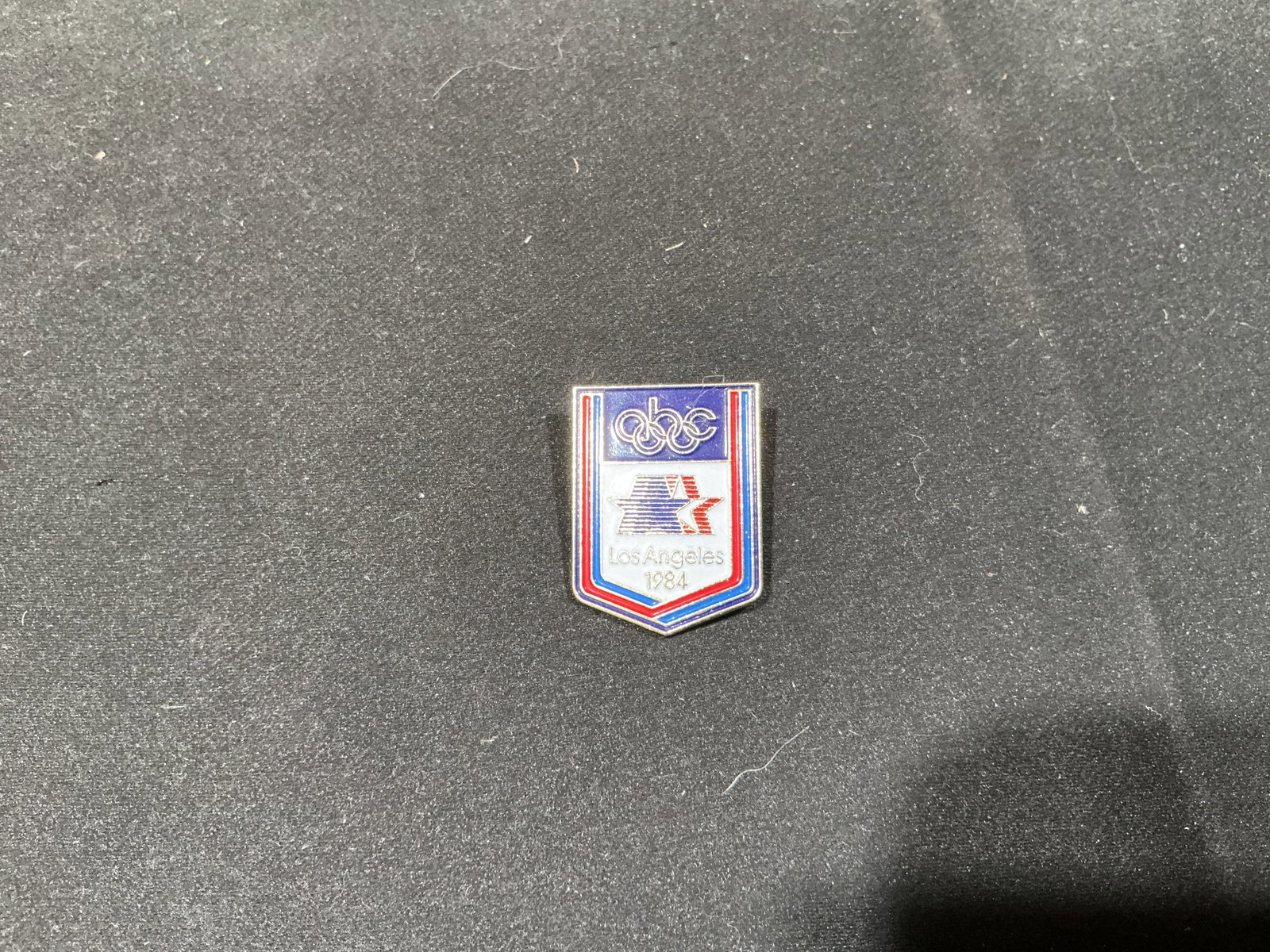 1984 Los Angeles Olympics ABC Television Pin: Created for the 1984 Summer Olympics in Los Angeles, this shield-shaped enamel pin combines the Olympic rings with the red, white, and blue Los Angeles 1984 star-in-motion emblem and ABC branding.