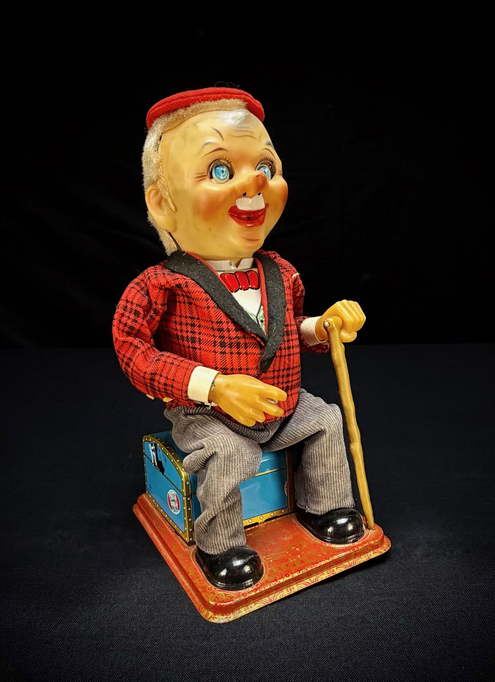 Vintage Nomura (TN) Smoking McGregor Tin Toy: This is a vintage battery-operated smoking McGregor toy by Nomura (TN) of Japan. The figure is dressed in a red plaid jacket with grey trousers and a red beret, seated on a tin lithograph suitcase whi
