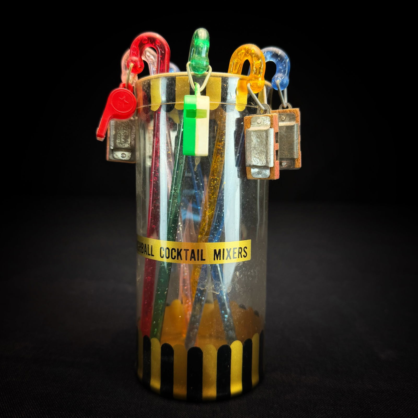 Vintage Highball Cocktail Mixers with Charms"Whistle for Another": This is a set of vintage Highball Cocktail Mixers in their original cylindrical packaging. The set includes multiple translucent plastic stirrers in various colors, each with glitter suspended inside.