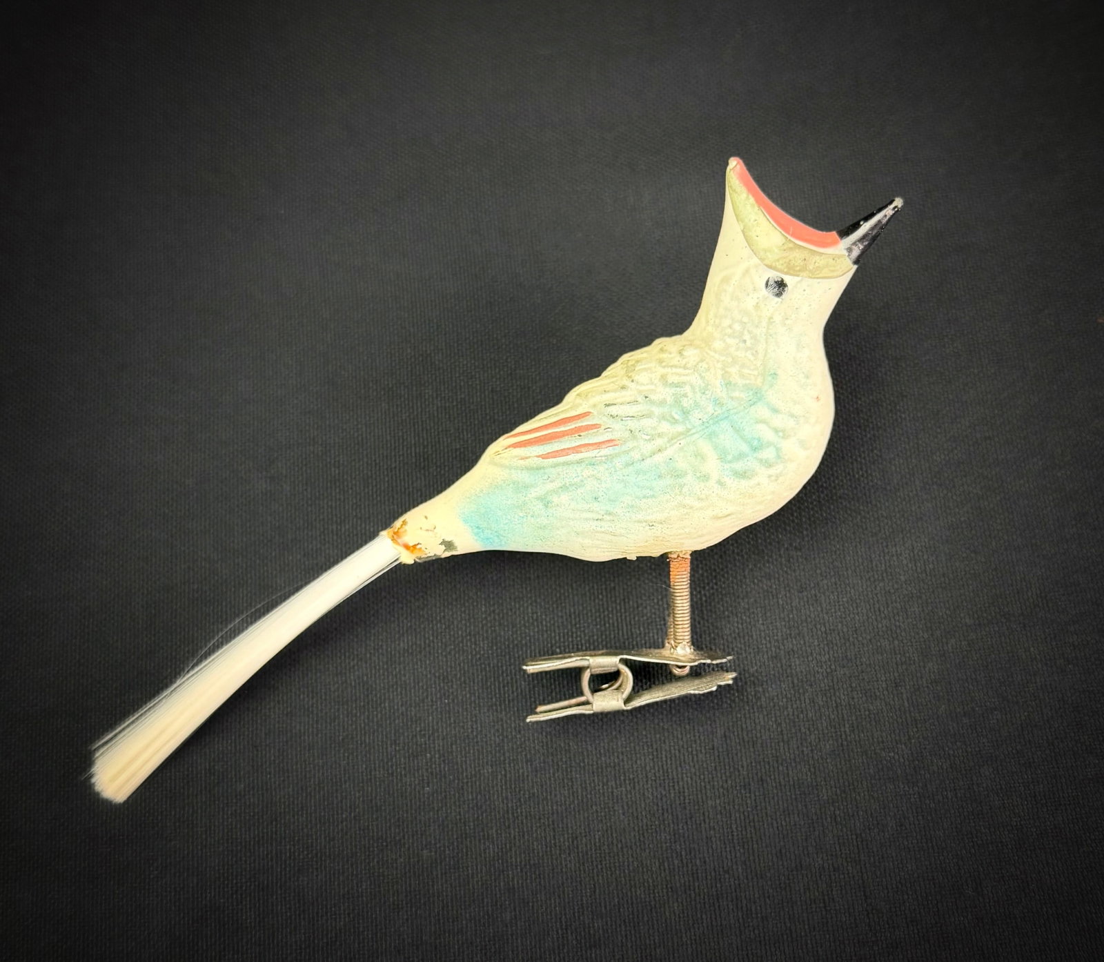 Antique German Glass Bird Ornament: An antique German clip-on Christmas ornament in the form of a bird. The piece features a molded glass body with painted details, a spun glass tail, and wire legs on a metal clip. The bird is depicted