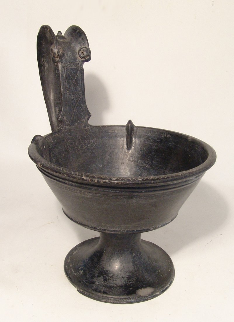 A large and impressive Etruscan bucchero kyathos: A large and impressive Etruscan bucchero kyathos, c. 560 – 520 BC, on a stemmed foot with large flanged handle adorned with two frontal ram heads in relief and surmounted by a nipple. The front of t