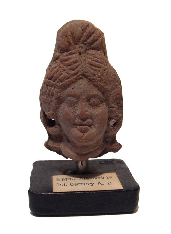 An Egyptian terracotta head of a woman, Alexandria: An Egyptian terracotta head of a woman, Alexandria, 1st Century AD, her features nicely detailed, hair styled in ridges with a coiled plait atop her hair, ears pierced for earrings in another substanc