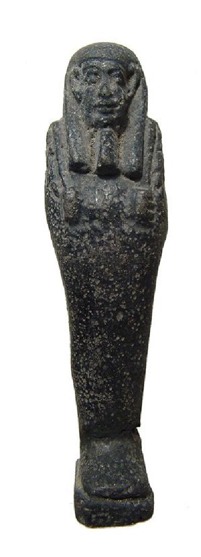 An Egyptian faience ushabti with black glaze, Late: An Egyptian faience ushabti with black glaze, Late Period, c. 664 - 30 BC, depicted mummiform and standing on integral base, wearing tripartite wig and false beard, arms folded at chest, hands holding