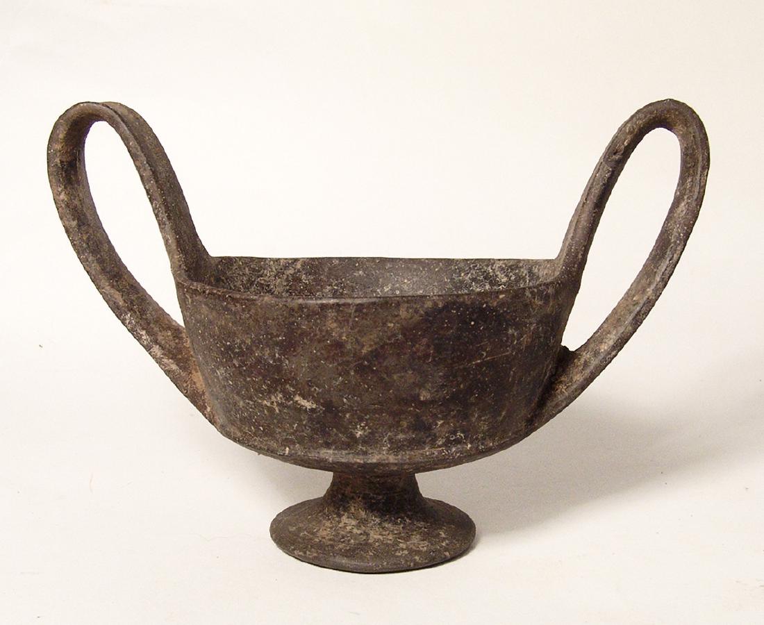 An Etruscan footed kantharos: An Etruscan footed kantharos, c. 625 - 550 BC, the bowl with ridged carination and lightly flared walls, low pedestal foot and a pair of tall loop handles. H: 4 3/4 in (12cm). Light surfaces deposits 