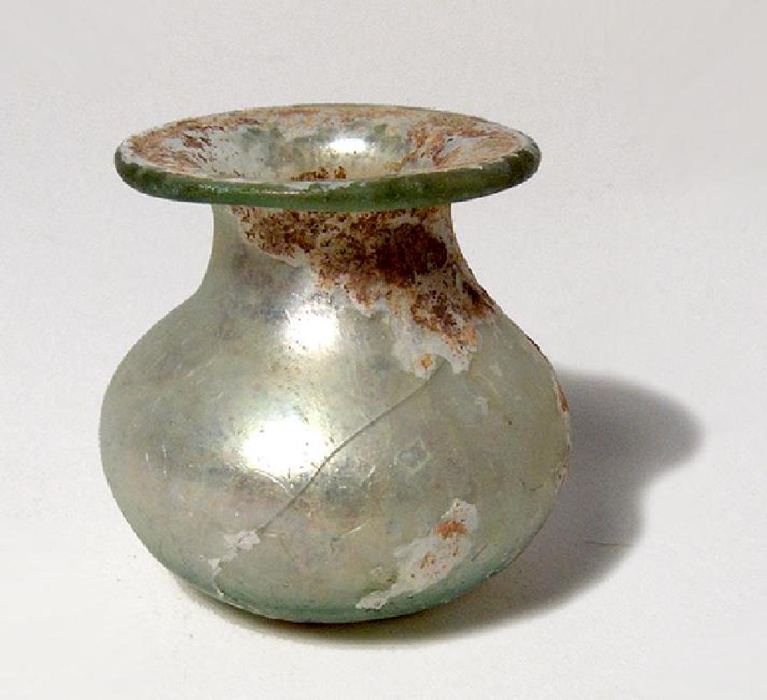 A squat Roman green glass jar: A squat Roman green glass jar, c. 2nd - 3rd Century AD, the ovoid body with flattened base, short neck with widely flared mouth with inward folded rim. H: 2 in (5.1cm). Light mineral deposits and irid