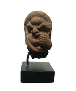 A Romano-Egyptian terracotta head of a grotesque: A Romano-Egyptian terracotta head of a grotesque, c. 1st Century BC/AD, possibly a comic actor, wearing a rolled hair fillet, which sits above a brow protuberance, his left forefingers in one side of