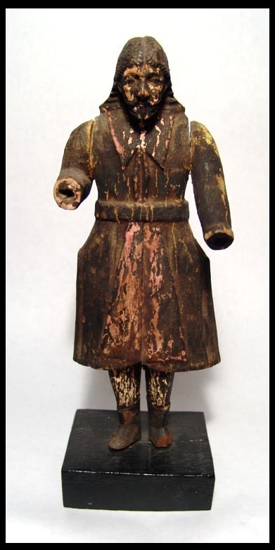Nice wooden male santos figure, mid 19th century: A nice wooden male santos figure, mid 19th century, wearing a long coat and belt with long hair and boots. Red and yellow pigment visible under black sooty deposits. Body carved from a single piece of