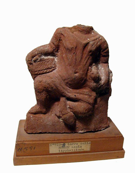 Terracotta figure of an Ithyphallic male: A terracotta figure of an Ithyphallic male, Ptolemaic Egypt, c. 2nd - 1st Century AD. Nicely molded, with head now lost. The figure rests on his knee and holds a cat and a basket. Traces of white dres
