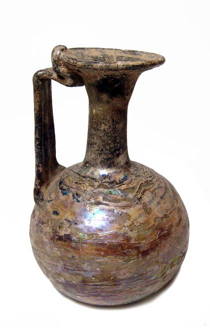 Vividly iridescent Roman glass bottle: A vividly iridescent Roman glass bottle, c. 3rd-4th Century AD, with single strap handle adorned with three vertical ridges. The body is round with indented base, the neck narrow and mouth flared wide