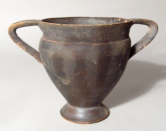 An attractive Greek black-ware cup: An attractive Greek black-ware cup, c. 4th - 3rd Century BC, the body tapering to a flared foot, the vertical rim offset by a groove above the shoulder and two triangular-shaped handles. H: 4" (10.1 c