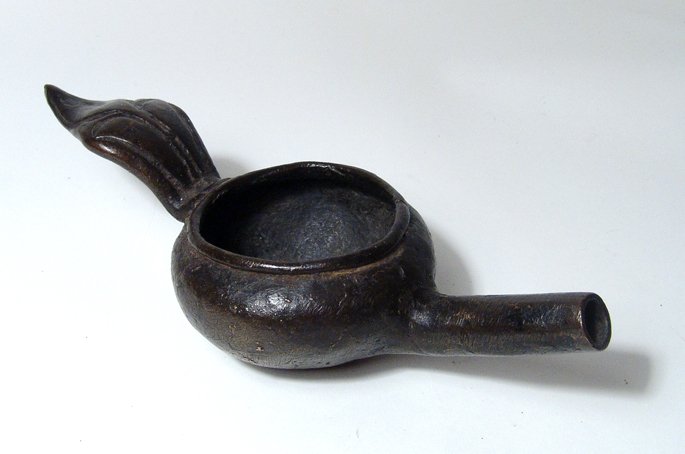 Near Eastern antique bronze lamp: A Near Eastern antique bronze spouted lamp, c. 19th Century, with long tubular spout, the body, round with wide mouth and rounded rim, the handle in the form of a leaf. L: 8 in (20.4 cm). Intact, warm