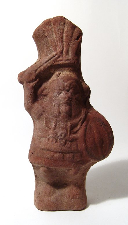 Egyptian terracotta figure of the god Bes: An Egyptian terracotta figure of the god Bes, Ptolemaic, 3rd - 2nd Century BC, armed, his sword raised overhead and holding an oval Gaulish shield at his left side. He wears a high feathered crown as 