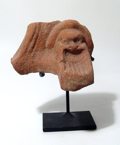A terracotta pot-support with mask of Papposeilenos: A terracotta pot-support with mask of Papposeilenos, Ptolemaic Period, late 3rd – Mid-1st Century BC, with long striated beard, within an arched panel. The back is plain, the edge of the fire bowl s