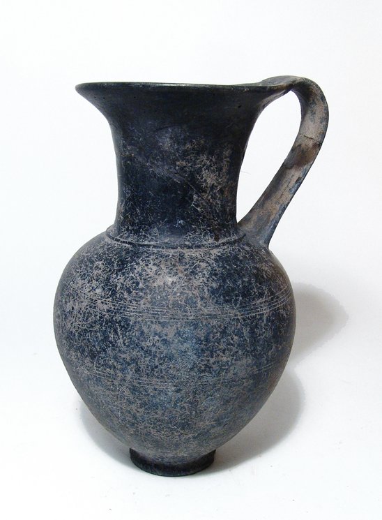 A large Etruscan Bucchero olpe: A large Etruscan Bucchero olpe, c. 6th Century BC, the tapering body decorated with incised banding. A nice example with scattered mineral deposits. H: 9 in (22.7cm). Ex South German private collectio