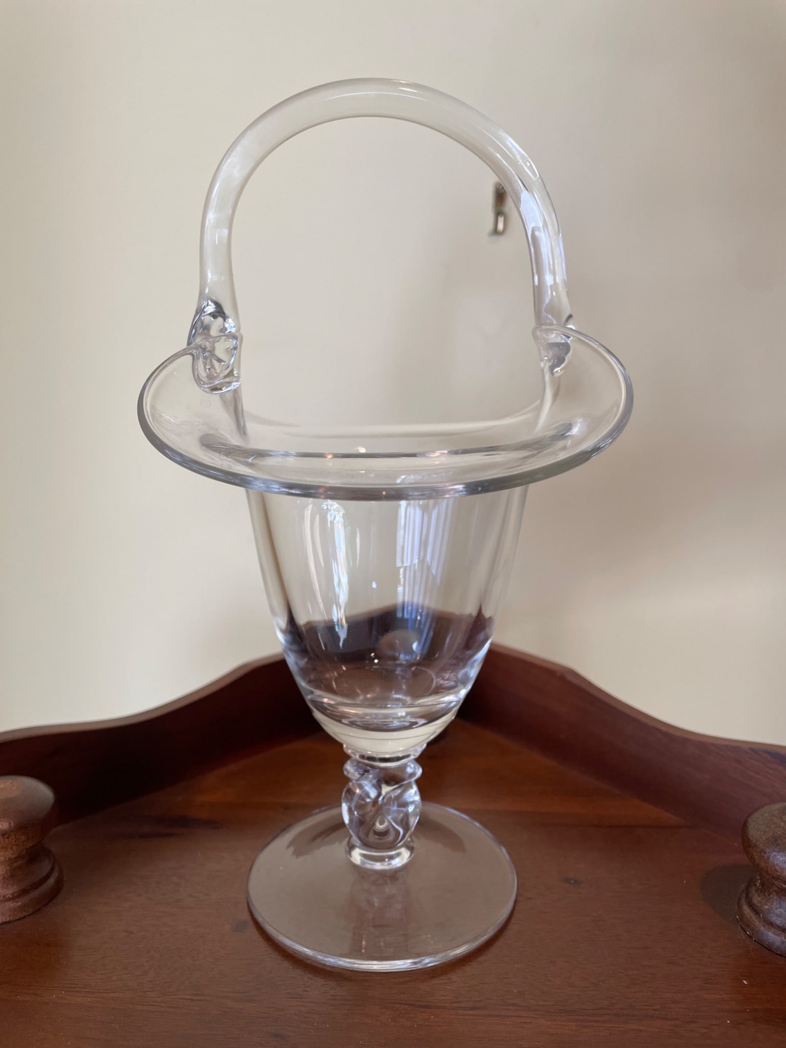 Clear Glass Pedestal Basket With Twisted Stem and Applied Handle: This is a clear glass pedestal basket with a twisted stem design and an applied handle. The item features smooth surfaces with a slightly flared rim and stands on a round, flat base. The handle is