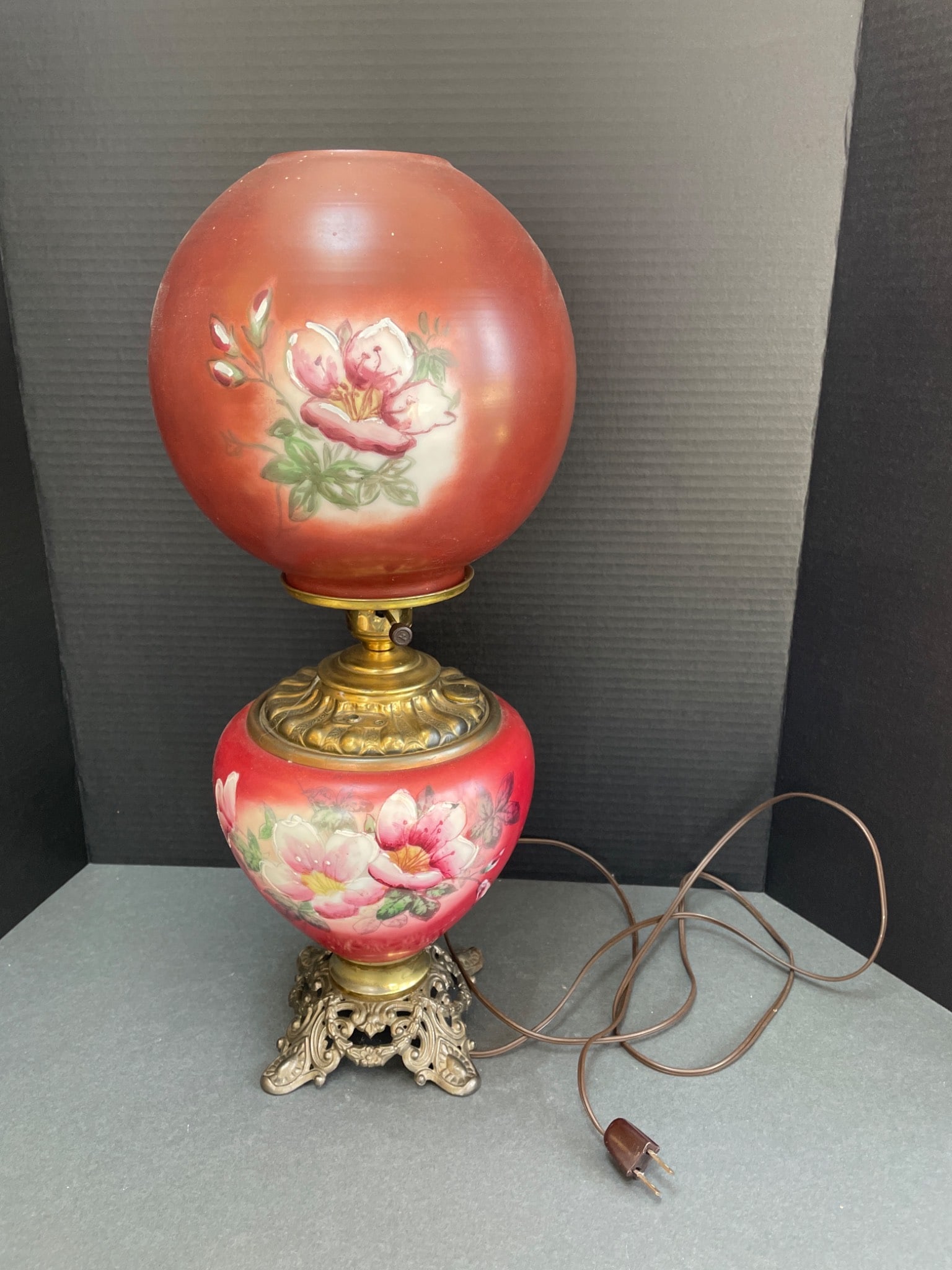 Electric Lamp With Hand-Painted Floral Design and Ornate Metal Base: Electric lamp featuring a hand-painted floral design on its glass globe and base. It includes an ornate metal base and a matching red-toned glass globe. The item includes a power cord with plug and ap