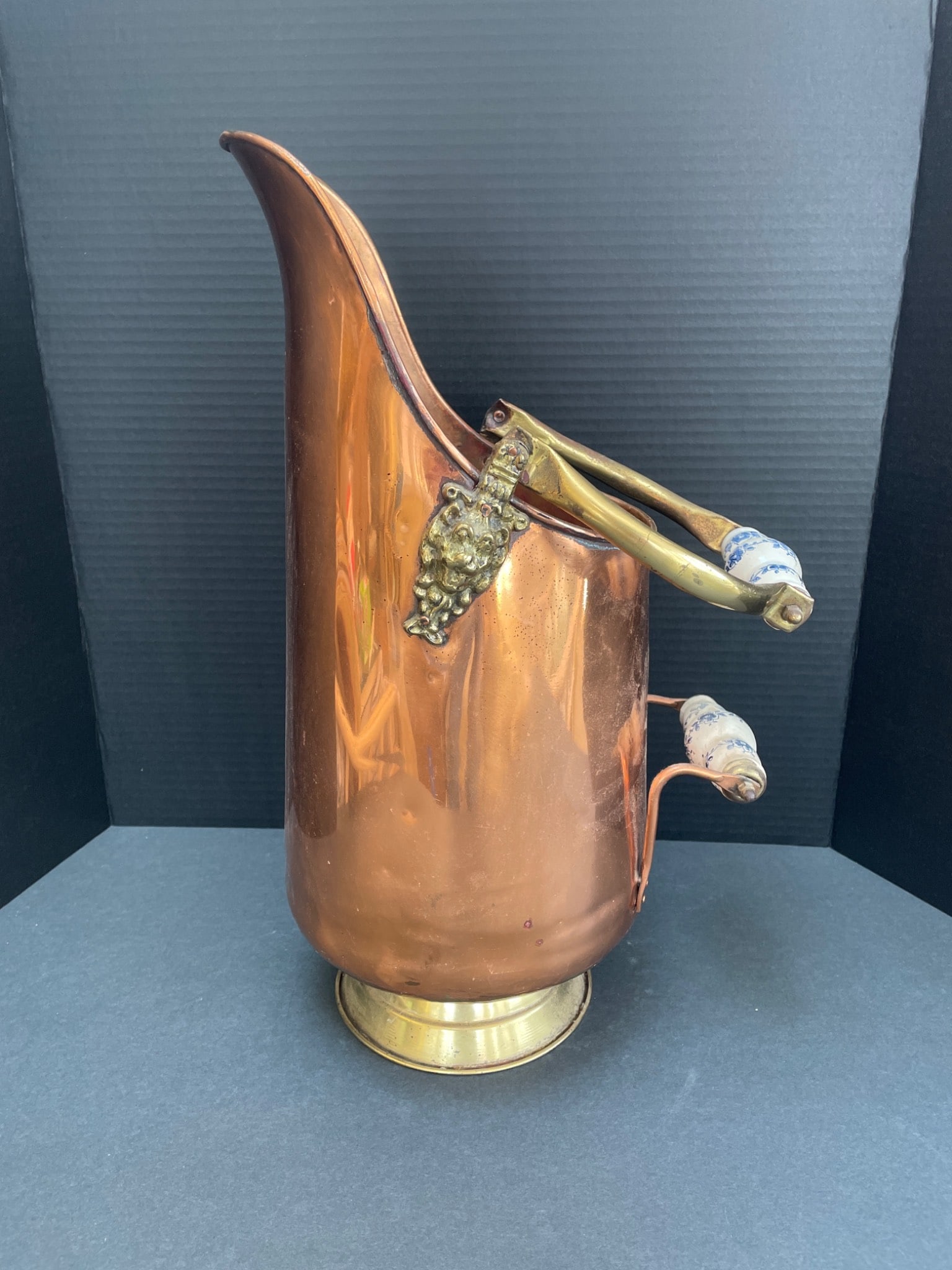 Copper and Brass Pitcher with Porcelain Handles: A copper and brass pitcher featuring decorative porcelain handles with blue floral motifs. The pitcher is designed with a curved spout and fixed handles secured by decorative brass brackets. It has a