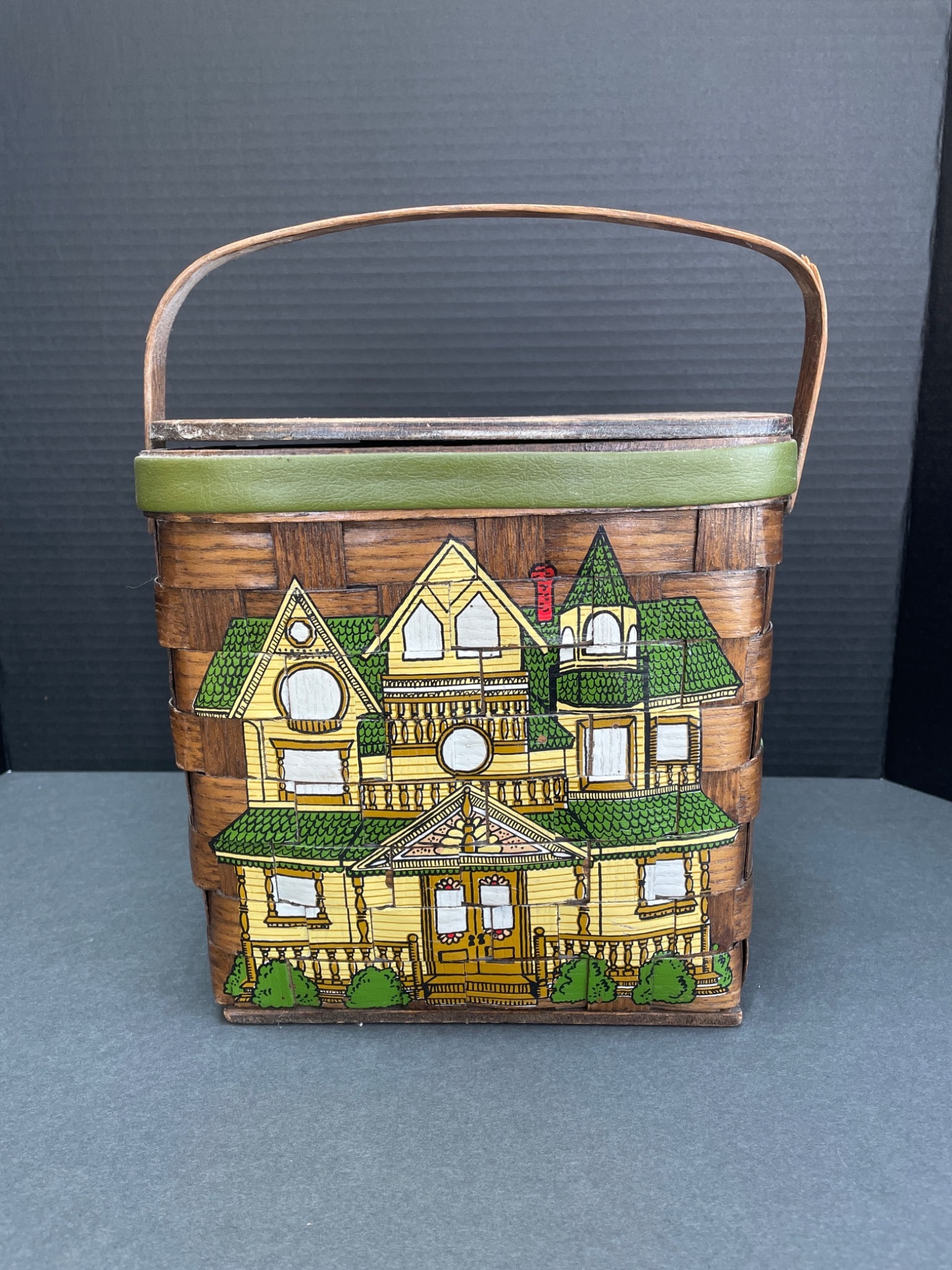 Rectangular Wooden Basket With Hinged Lid and Painted House Design: Rectangular woven wooden basket featuring painted decorative motifs, including a house and tree. The basket has a hinged wooden lid with lining and a reflective insert on the interior of the lid. Hand