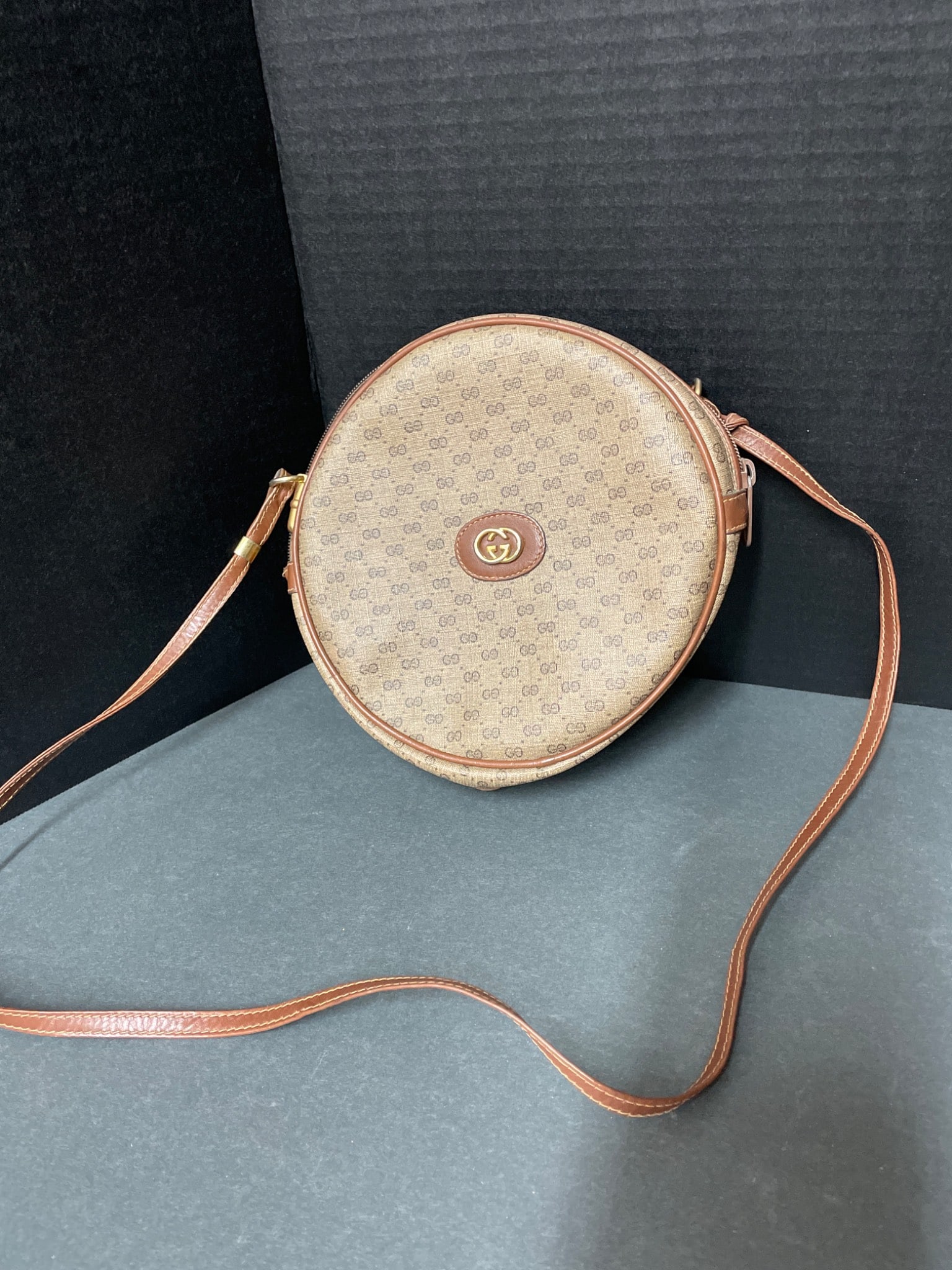 Round Crossbody Bag With Monogram Details and Leather Trim: This is a round crossbody bag featuring monogram detailing and leather accents. The bag includes a zip-around closure, an adjustable leather strap, and an interior zippered pocket. The logo hardware