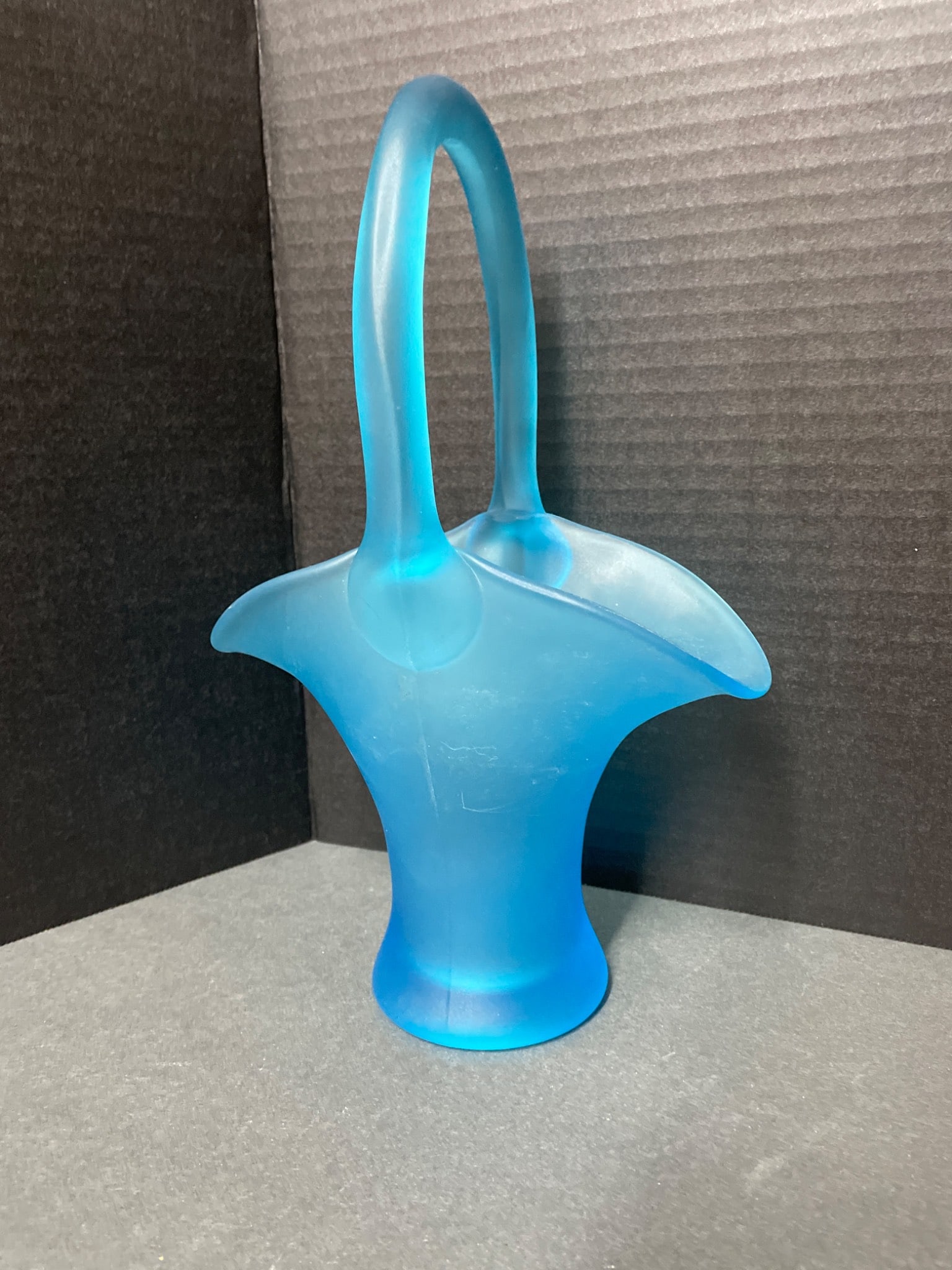 10 3/4-Inch Tall Frosted Blue Glass Basket With Handle: This is a frosted blue glass basket, measuring 10 3/4 inches tall. The piece features a wide flared rim, an integrated handle with a curved design, and a flat base for stability. The glass exhibits a