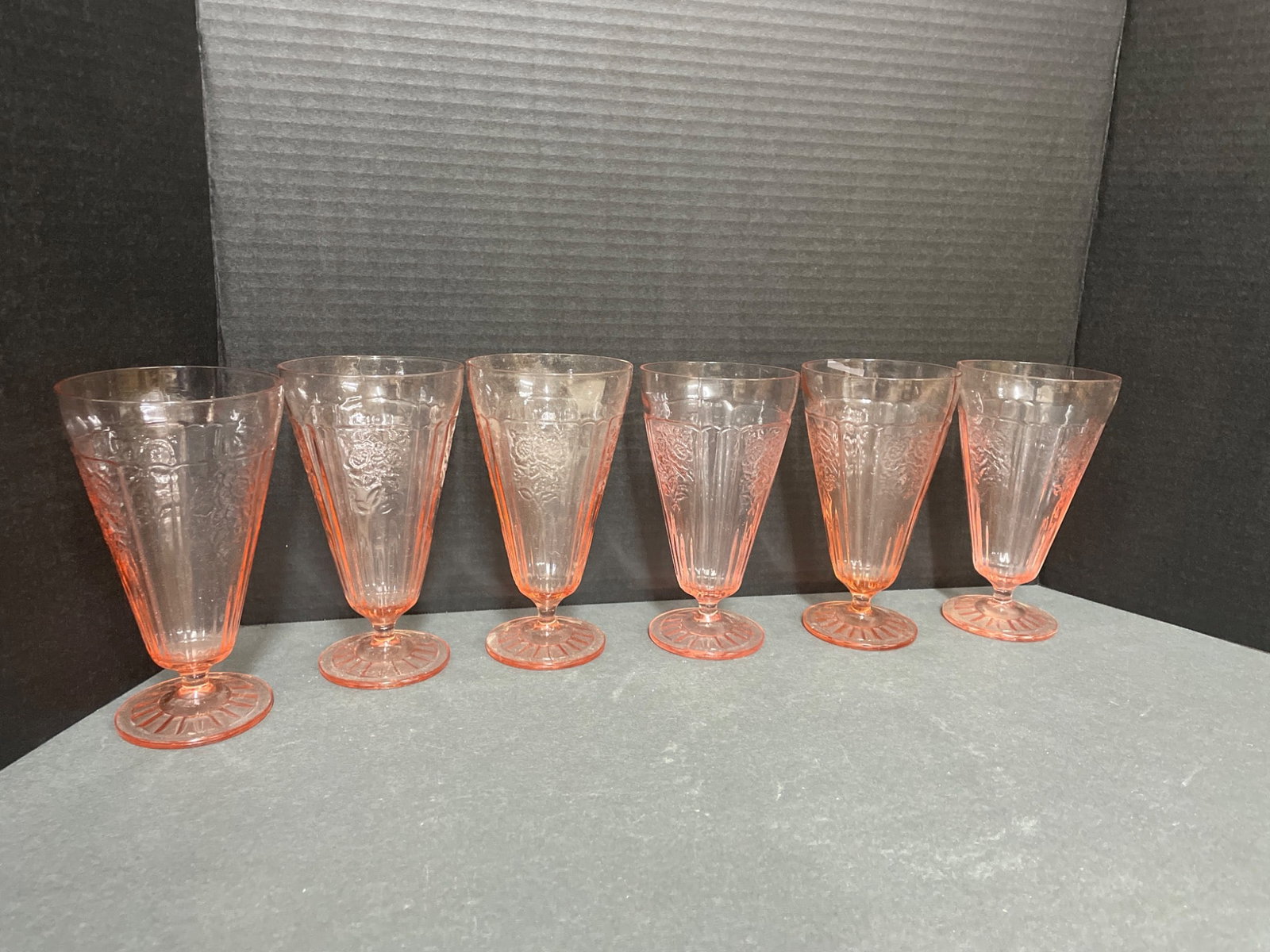 6 Pink Depression Glass Footed Tumblers with Floral Etching, 8 Inches: 6 matching footed tumblers in pink Depression glass. Each tumbler measures approximately 8 inches tall. Features vertical panel detailing with floral etched decorative designs. Constructed from