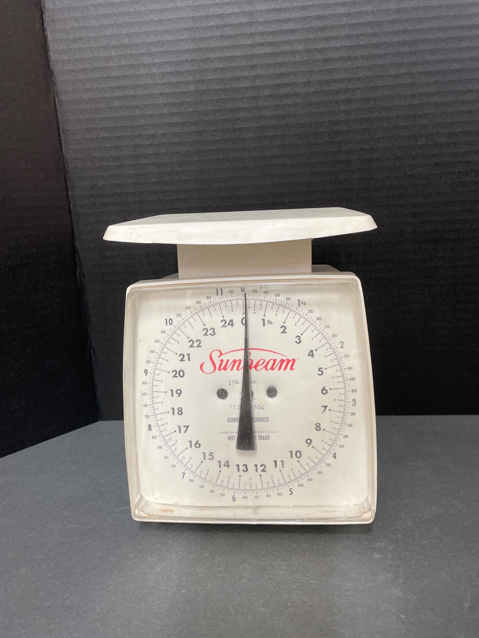 Sunbeam 25 lb / 12 kg Mechanical Dial Scale: This Sunbeam mechanical dial scale features a capacity of 25 lb (12 kg) with clear dual metric and imperial measurement markings. Model details are not clearly visible in the images. The unit is