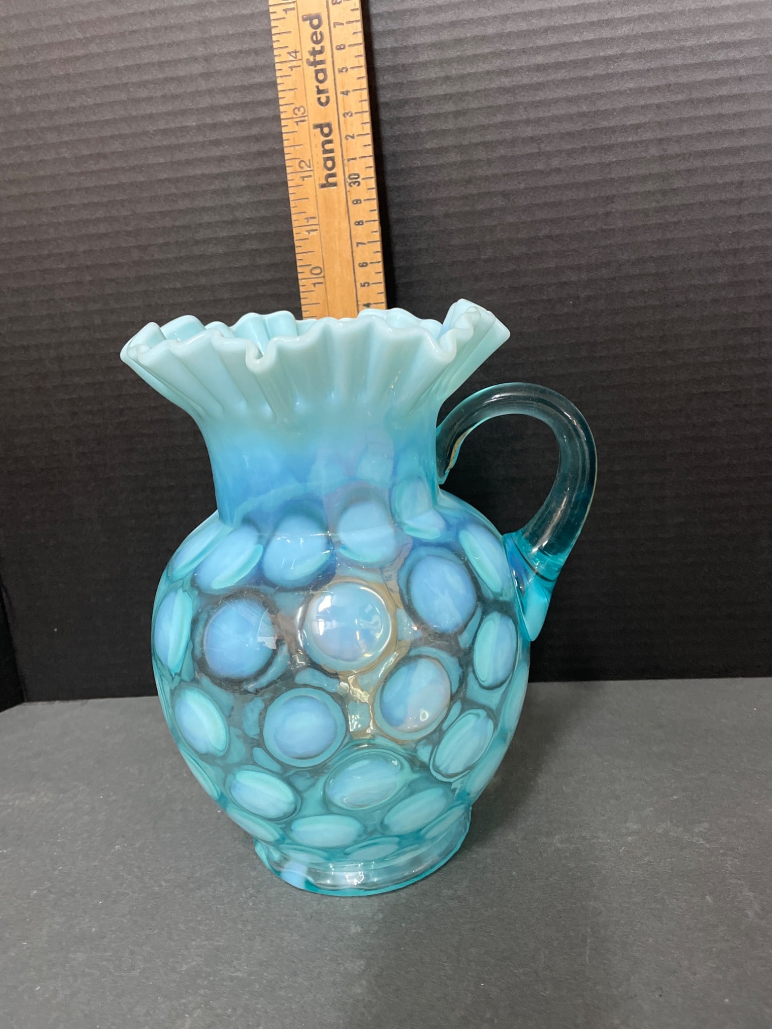 Blue Opalescent Glass Pitcher With Fenton Hobnail Design: Blue opalescent glass pitcher with a ruffled rim and hand-applied handle. Features a hobnail pattern consistent with Fenton's design characteristics. Dimensions are approximately 8 inches tall as